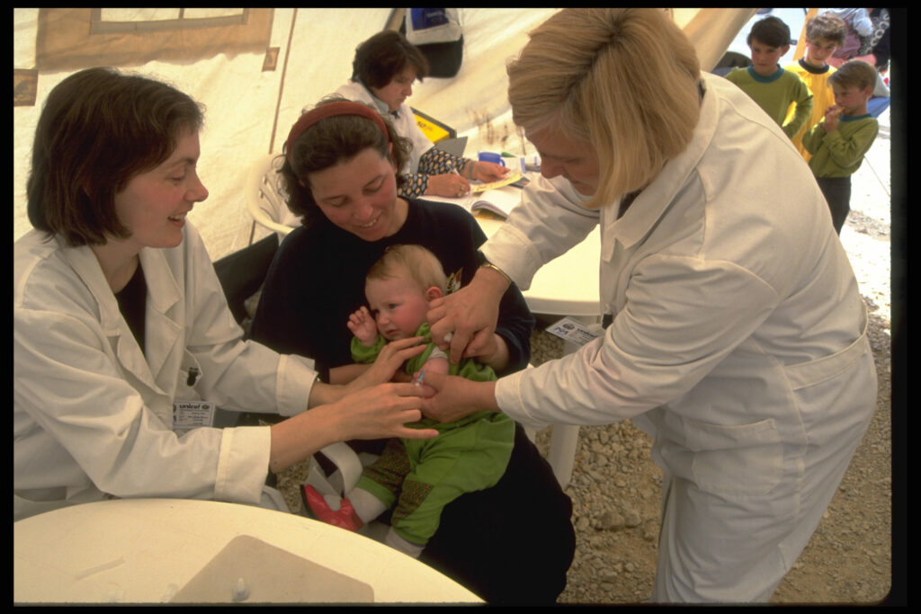 Een baby in de armen van een vrouw wordt gevaccineerd door een zorgmedewerker, terwijl een andere zorgmedewerker assisteert, in een door UNICEF ondersteunde kliniek in een tent in kamp Cegrane in het noordwesten van Macedonië. Cegrane bood tijdelijk onderdak aan ongeveer 40.000 mensen uit Kosovo en was het grootste vluchtelingenkamp van het land. Het kamp werd beheerd door de internationale hulporganisatie CARE. © UNICEF/UNI31684/Thomas