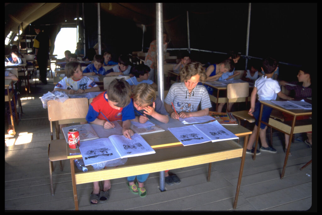 Kinderen uit Kosovo zitten aan tafels te werken in hun schriften in een ‘tentschool’ van UNICEF in het vluchtelingenkamp Cegrane in het noordwesten van Macedonië. Met meubilair en lesmateriaal van UNICEF bieden de tentscholen de kinderen de broodnodige stabiliteit en zorgen ze ervoor dat hun onderwijs kan doorgaan. Cegrane bood tijdelijk onderdak aan ongeveer 40.000 mensen uit Kosovo en was het grootste vluchtelingenkamp van het land. Het kamp werd beheerd door de internationale hulporganisatie CARE. © UNICEF/UNI31693/Thomas